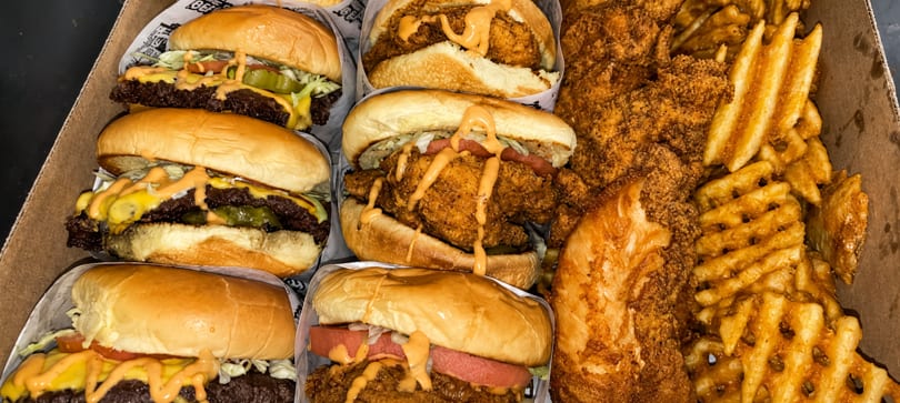 A cardboard box filled with six burgers, three beef and three fried chicken, alongside waffle fries.
